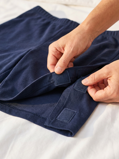 Caregiver opening velcro side panel on women's side-opening recovery shorts laid flat on bed [bedridden] | Aasra