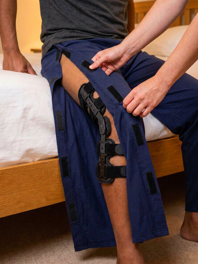 Caregiver helping man in dark charcoal tee with black hinged knee brace put on side-opening pyjamas [knee] | Aasra