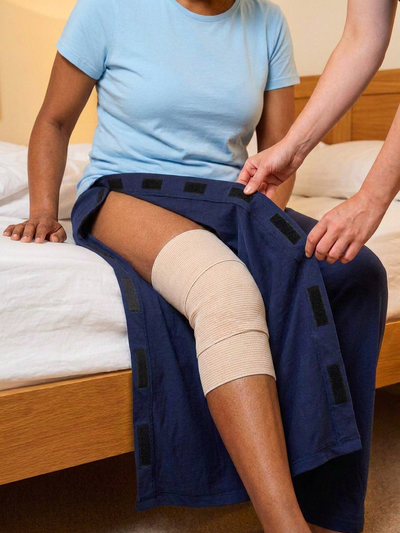 Caregiver helping woman in light blue top with beige bandage knee wrap put on side-opening pyjamas [knee] | Aasra